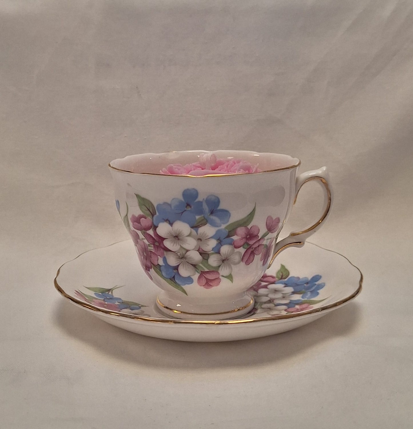 Side view of vintage floral teacup candle with blue, pink, and white flower design on bone china with gold trim.