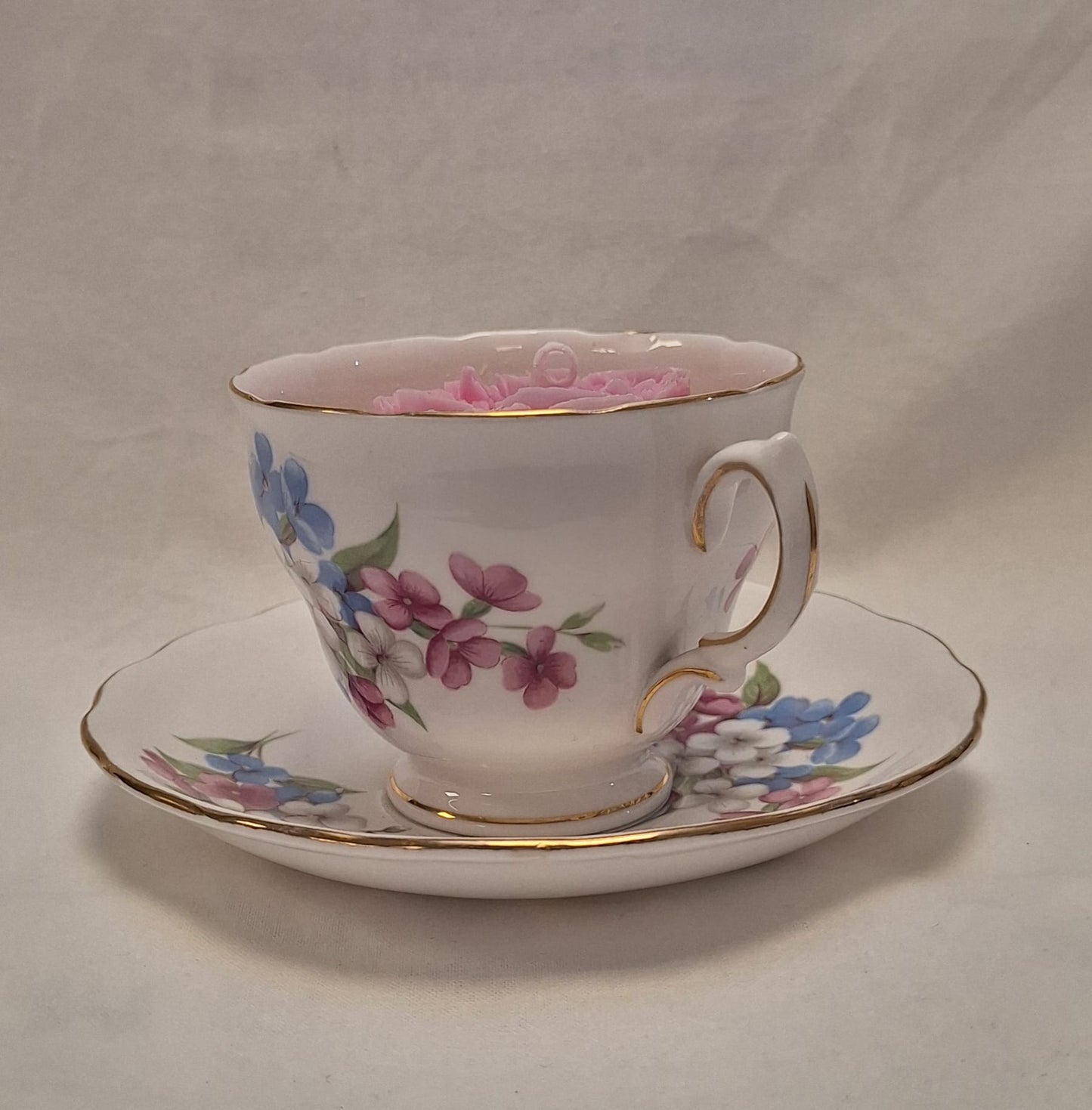 Side view of vintage floral teacup candle with blue, pink, and white flower design on bone china with gold trim.