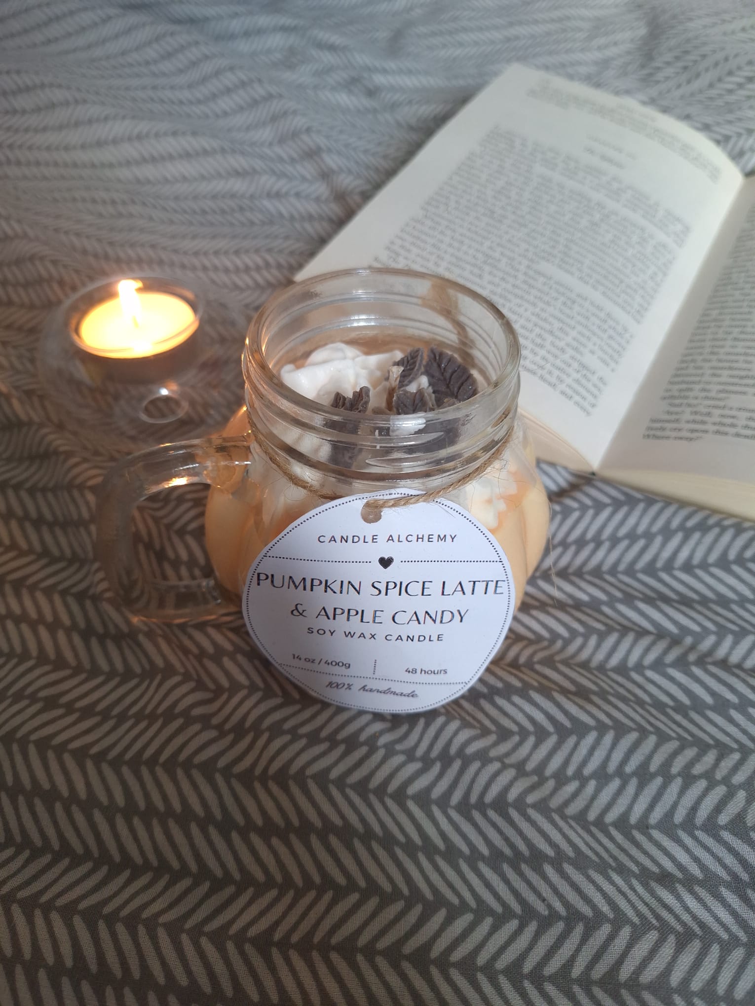 Pumpkin Spice Latte and Apple Candy soy candle in a mason jar with decorative toppings, next to a tealight and open book on a patterned blanket, perfect for autumn gifts and home décor.