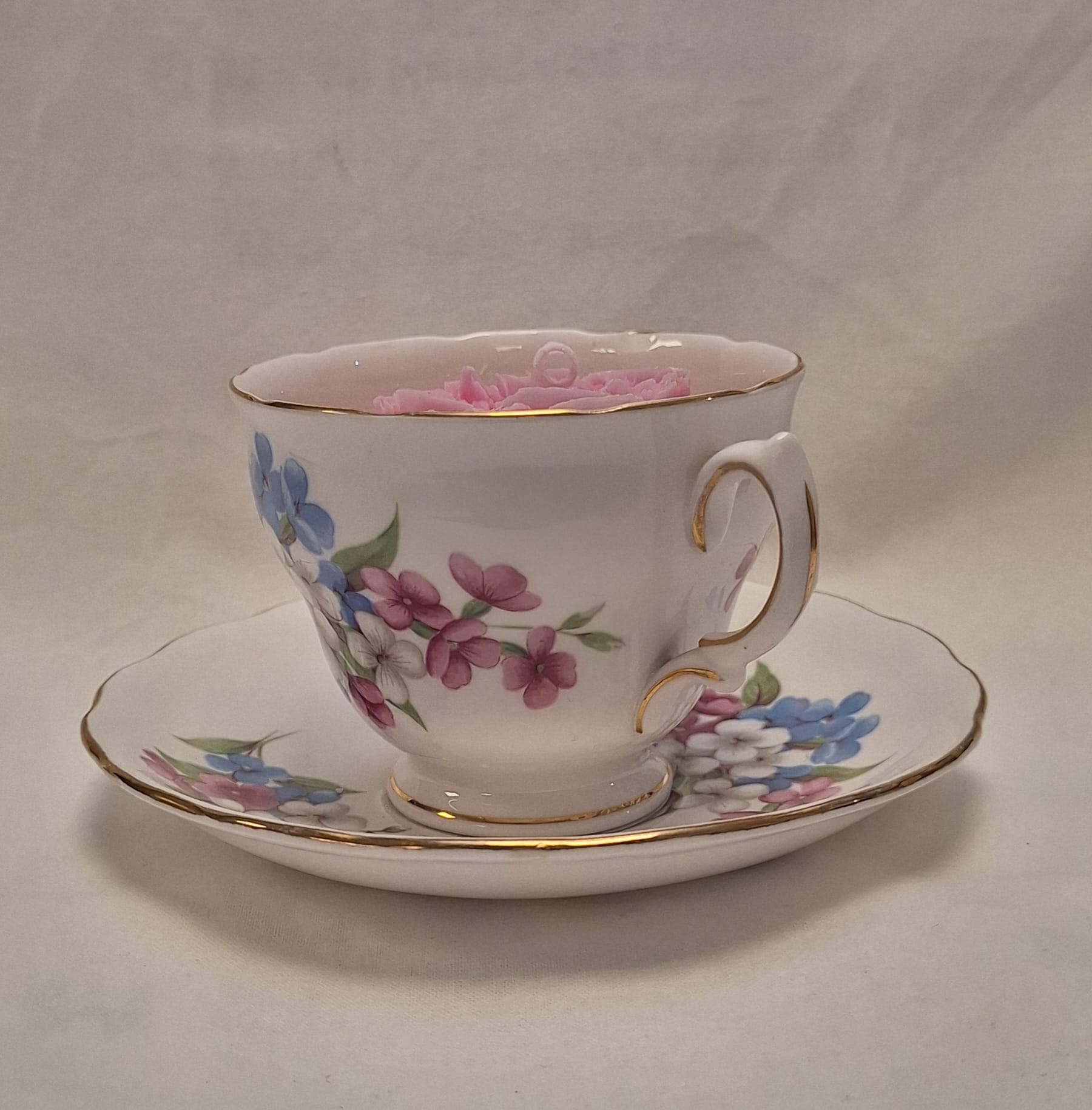 Side view of vintage floral teacup candle with blue, pink, and white flower design on bone china with gold trim.